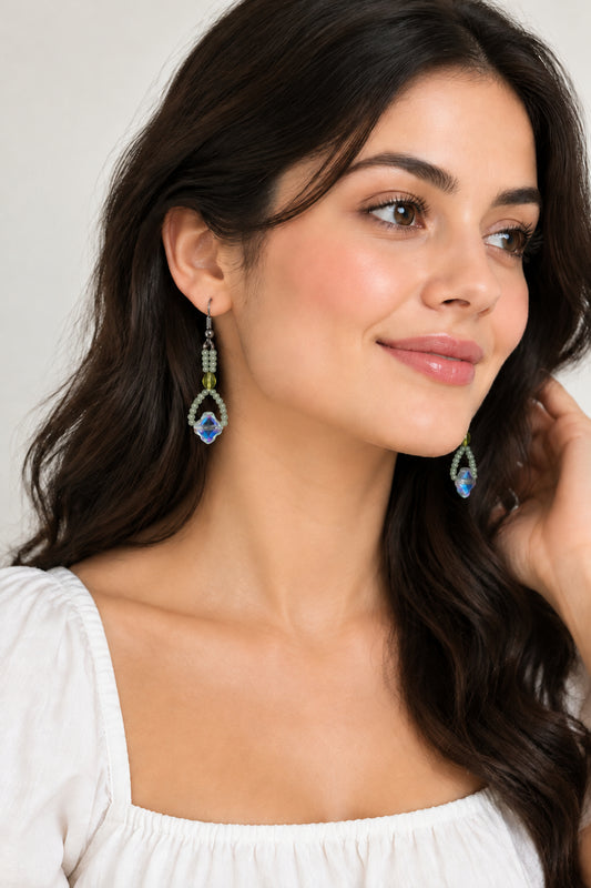 A pair of handmade beaded dangle earrings with silver fishhook wires, light green beads, and iridescent blue and purple star-shaped beads, displayed on a white faux fur background.